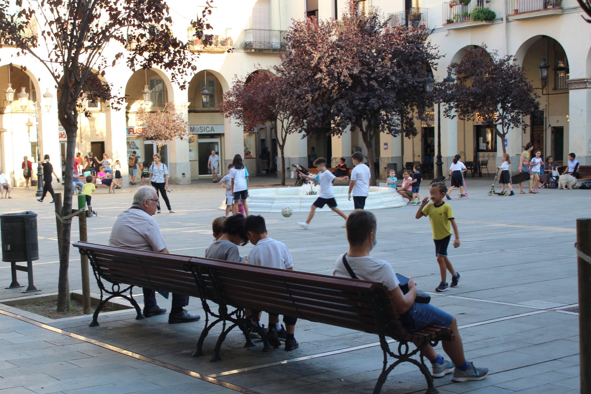 A la sortida de l'escola, molts infants juguen a pilota i corren per la plaça amb patinet mentre les famílies prenen un refresc al bar / Gabriel González