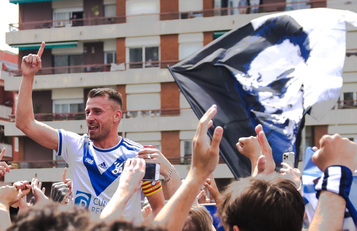 Àlex Cano és aixecat pels aficionats celebrant l'ascens de l'Europa amb la bandera de Manuel Cros, una altra llegenda de l'Europa, de fons | CE Europa